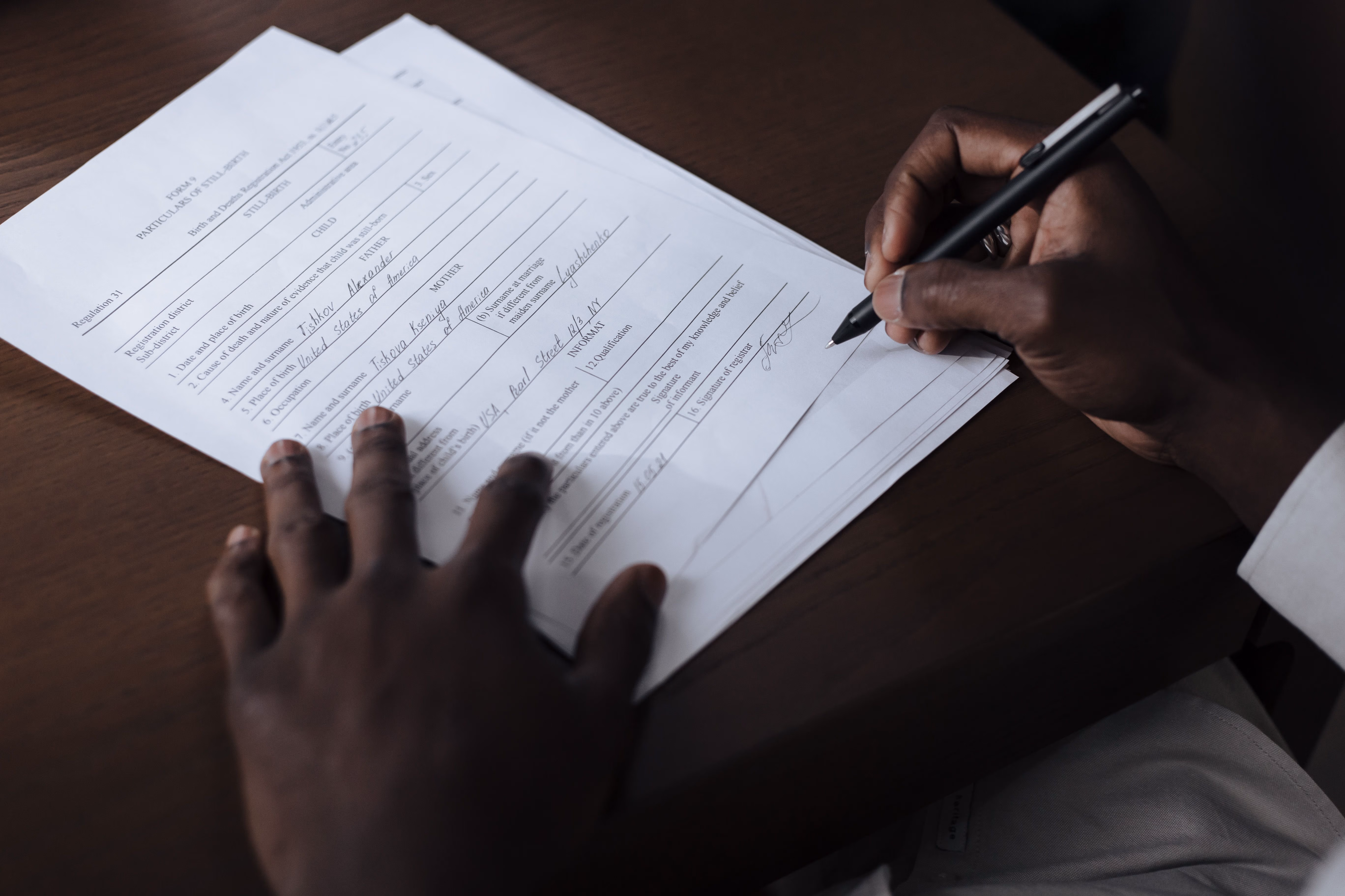 Person signing a stillbirth birth and death registration form on a wooden desk.